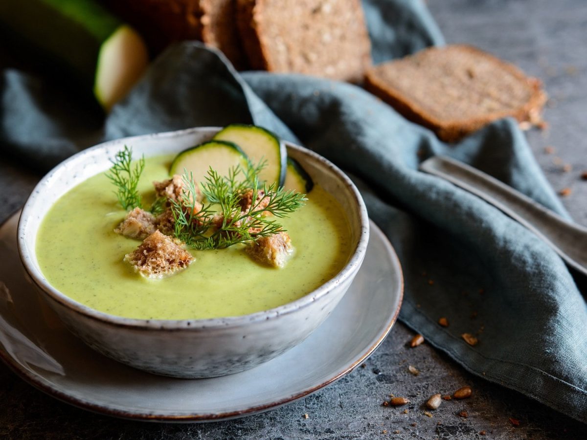 Eine Schüssel Zucchini-Spinat-Suppe mit Brotwürfeln, Zucchinischeiben und frischen Kräutern. Dahinter unscharf ein angeschnittenes Brot und zwei Zucchinis.