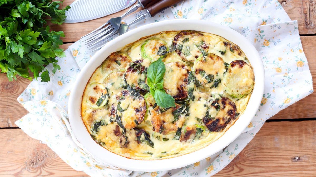 Zucchinigratin mit Kaesesauce in weißer Auflaufform auf Holz.