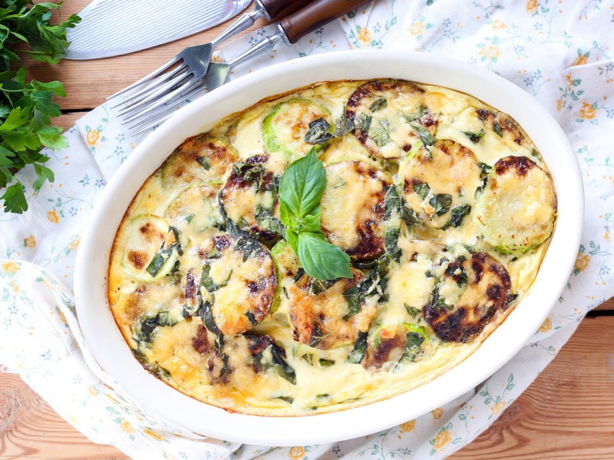 Zucchinigratin mit Kaesesauce in weißer Auflaufform auf Holz.