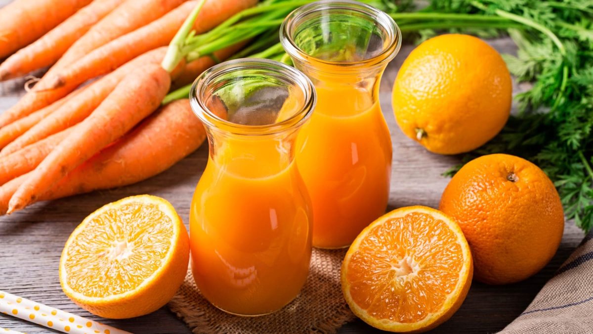 ACE-Saft selbst machen: Zwei Mini-Karaffen mit orangenem Saft, drum herum ganze und halbe Orangen und Möhren auf Holzuntergrund.