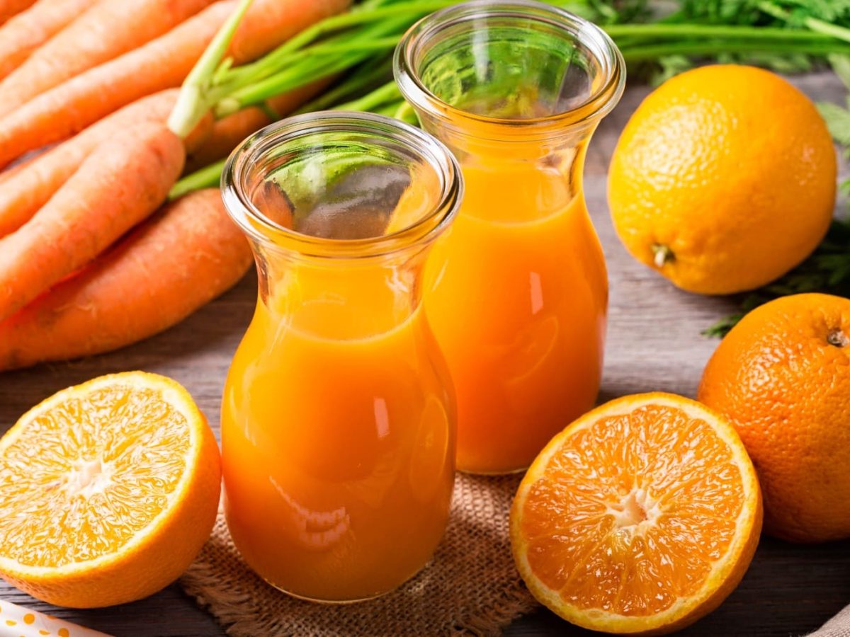 ACE-Saft selbst machen: Zwei Mini-Karaffen mit orangenem Saft, drum herum ganze und halbe Orangen und Möhren auf Holzuntergrund.