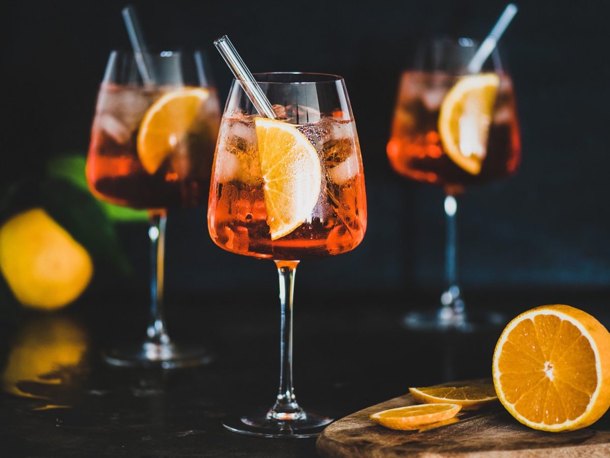 Drei Gläser alokoholfreier Aperol Spritz mit je einem Strohhalm und einer Orangenscheibe darin, daneben eine angeschnittene Orange.