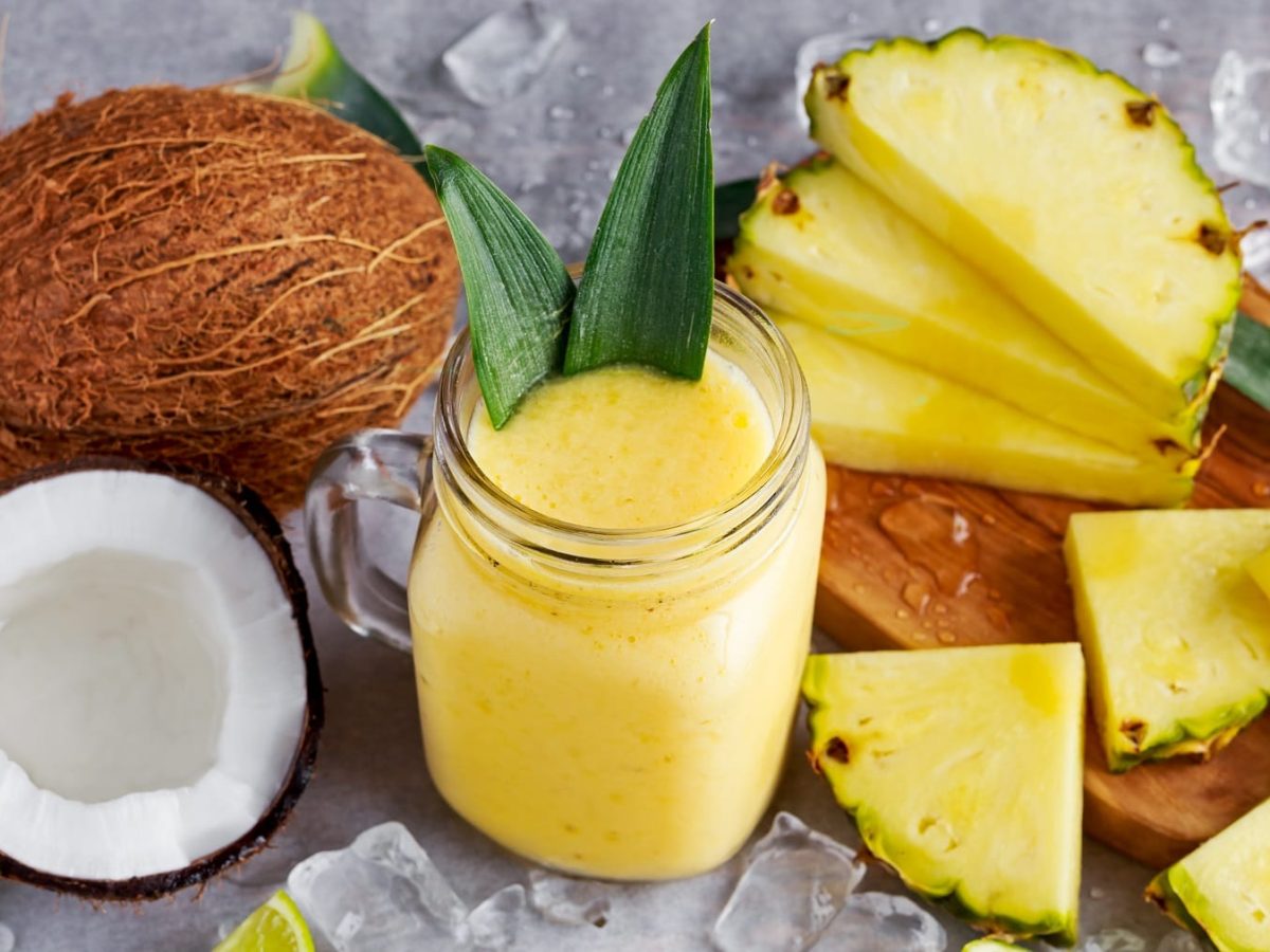 Ein Glas mit dem Ananas-Kokos-Smoothie auf einem grauen Tisch mit den beiden Früchten und Eiswürfeln.