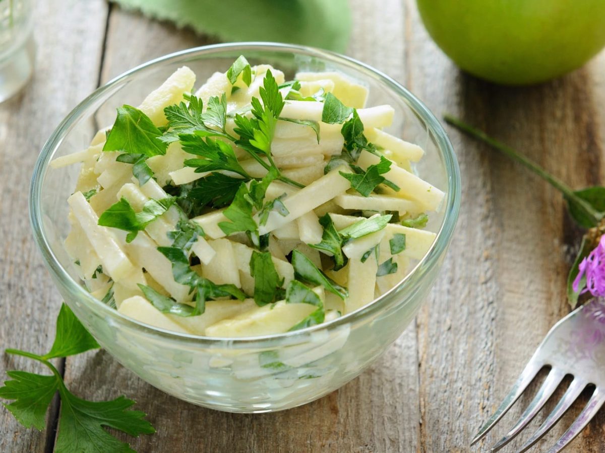 Eine Glasschale mit dem Apfel-Kohlrabisalat auf einem Holztisch mit Petersilie, lila Blüte und Granny Smith Apfel.