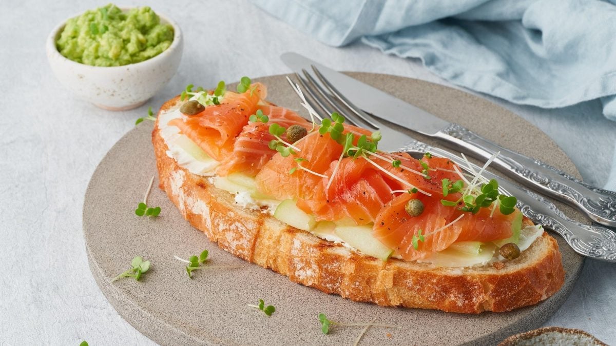 Draufsicht, schräg: ein Apfel-Lachs-Sandwich auf einem runden Holzbrett.