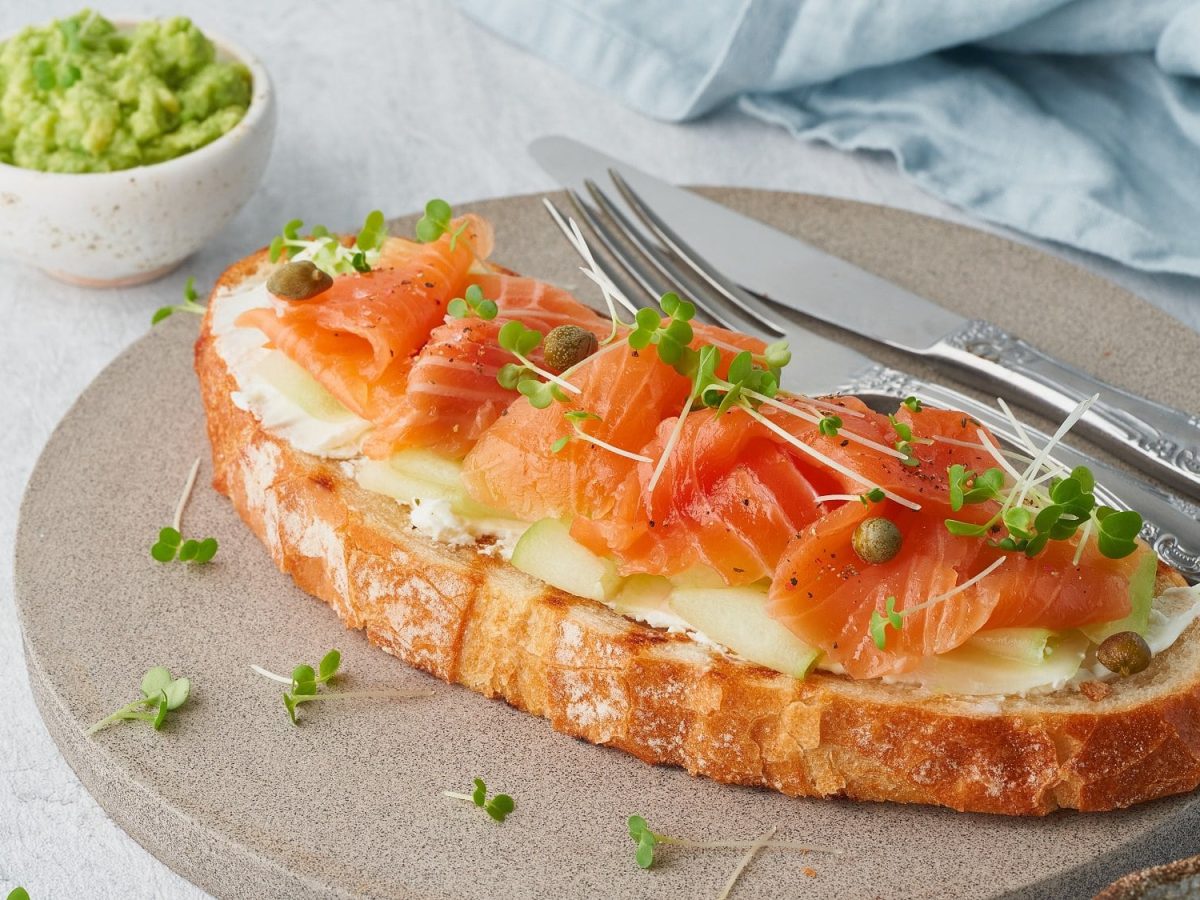 Draufsicht, schräg: ein Apfel-Lachs-Sandwich auf einem runden Holzbrett.