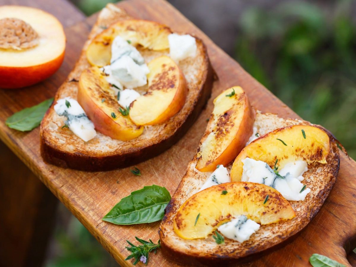 Draufsicht: Zwei Pfirsich-Bruschetta mit Gorgonzola auf einem Holzbrett. Daneben liebe Basilikumblätter und ein halber Pfirsich.