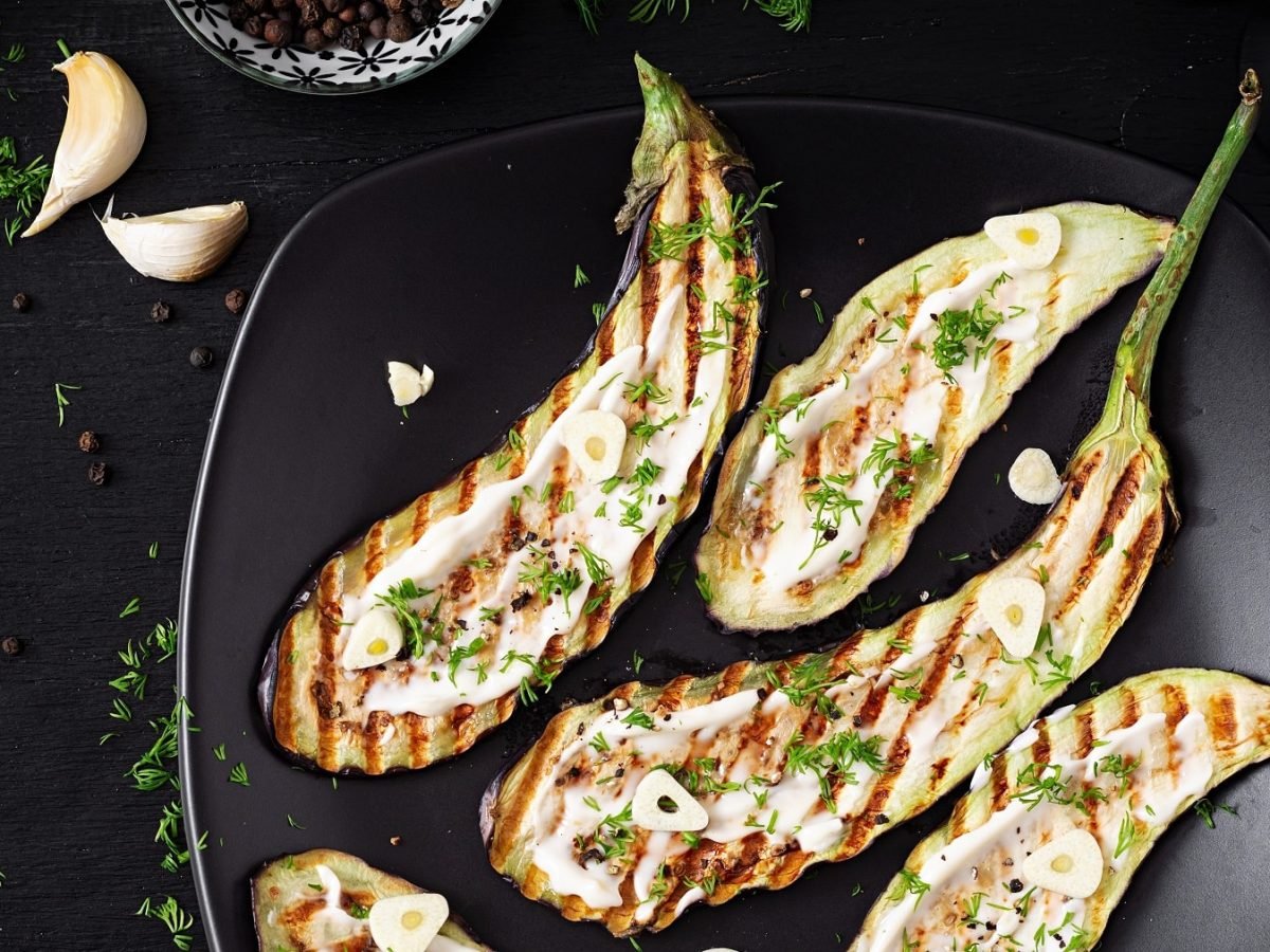 Einige Auberginenschieben mit Grillmuster auf einem schwarzen Teller mit Sesam-Joghurt und frischer Petersilie.