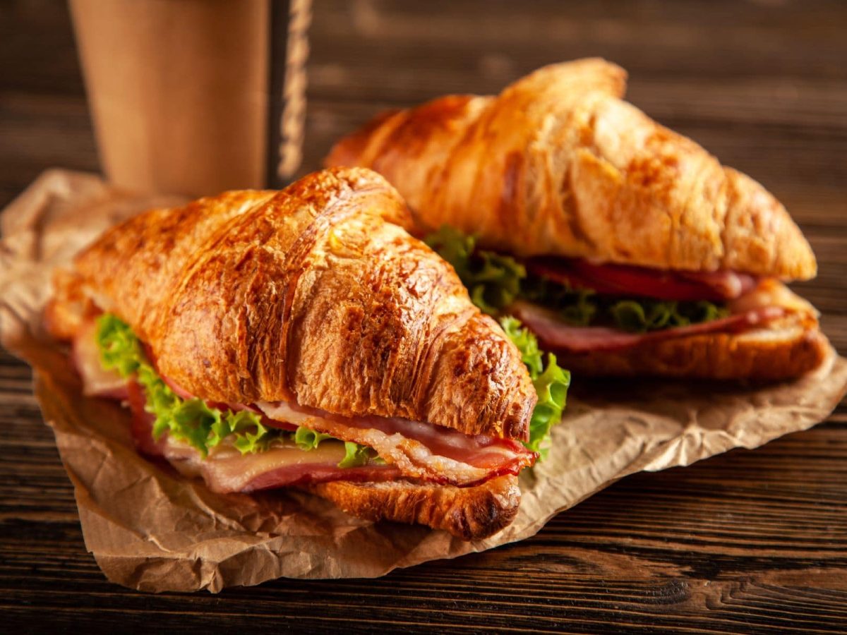 Zwei BLT-Croissant-Sandwiches auf Papier mit einem To-Go-Kaffeebecher auf Holz. Draufsicht.