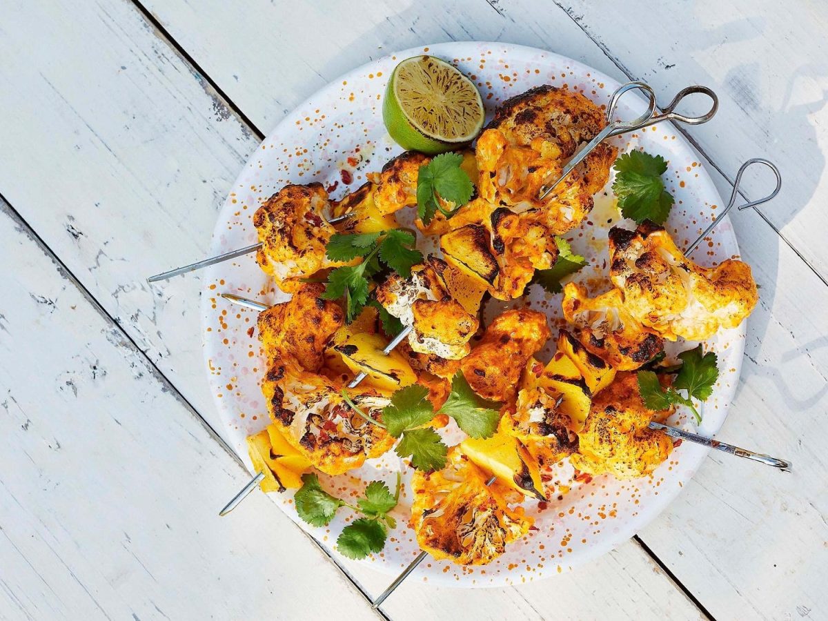 Blumenkohl-Mango-Spieße auf einem Teller mit Limetten und Koriander in der Draufsicht.