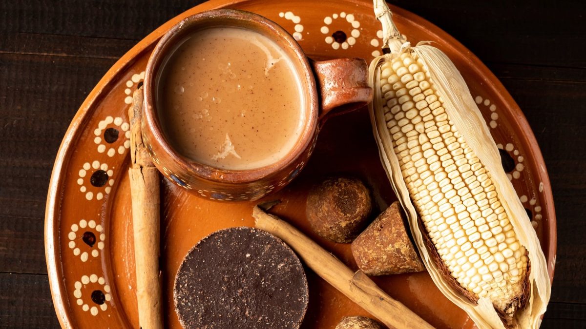 Ein brauner Teller mit einer Tasse Champurrado auf schwarzem Untergrund. Daneben Maiskolben, Rohrohrzucker und Zimt. Alles von oben fotografiert.