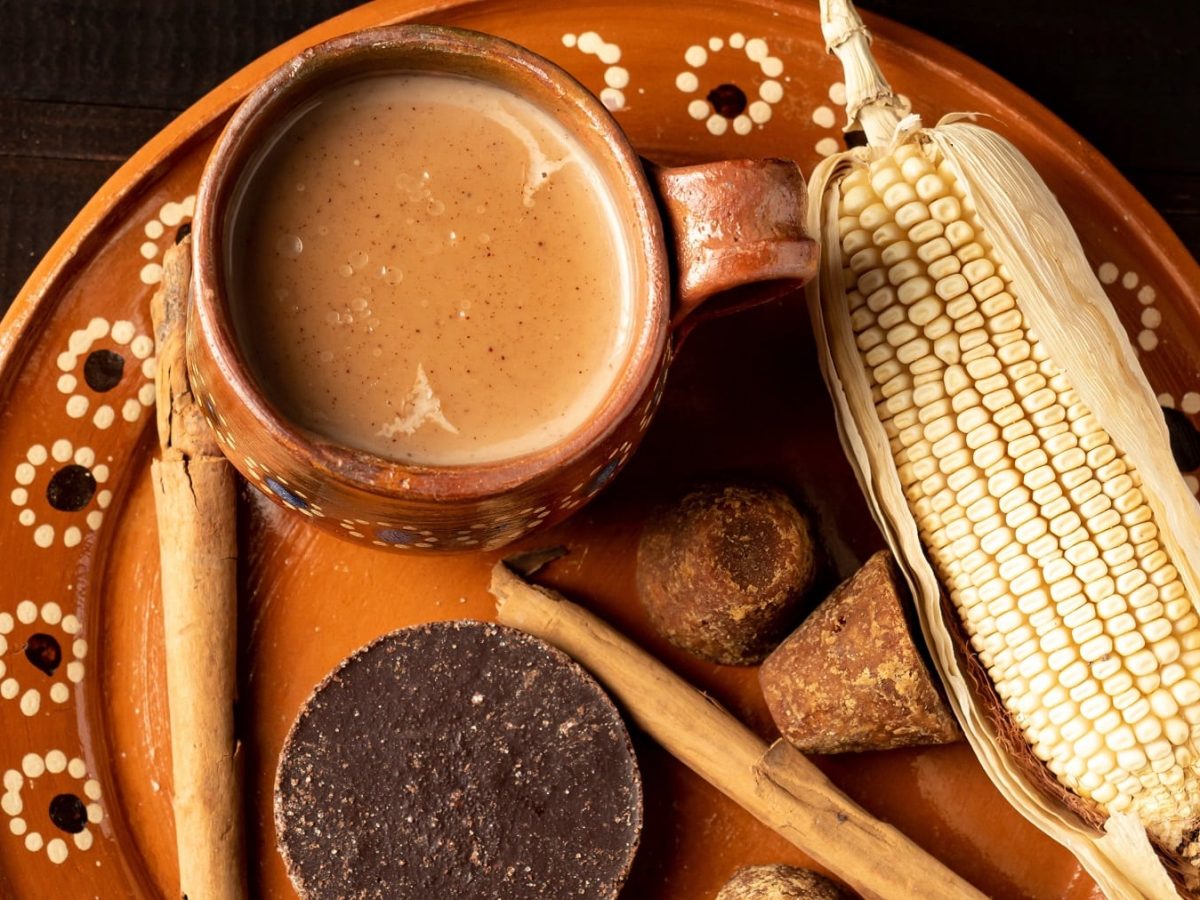 Ein brauner Teller mit einer Tasse Champurrado auf schwarzem Untergrund. Daneben Maiskolben, Rohrohrzucker und Zimt. Alles von oben fotografiert.