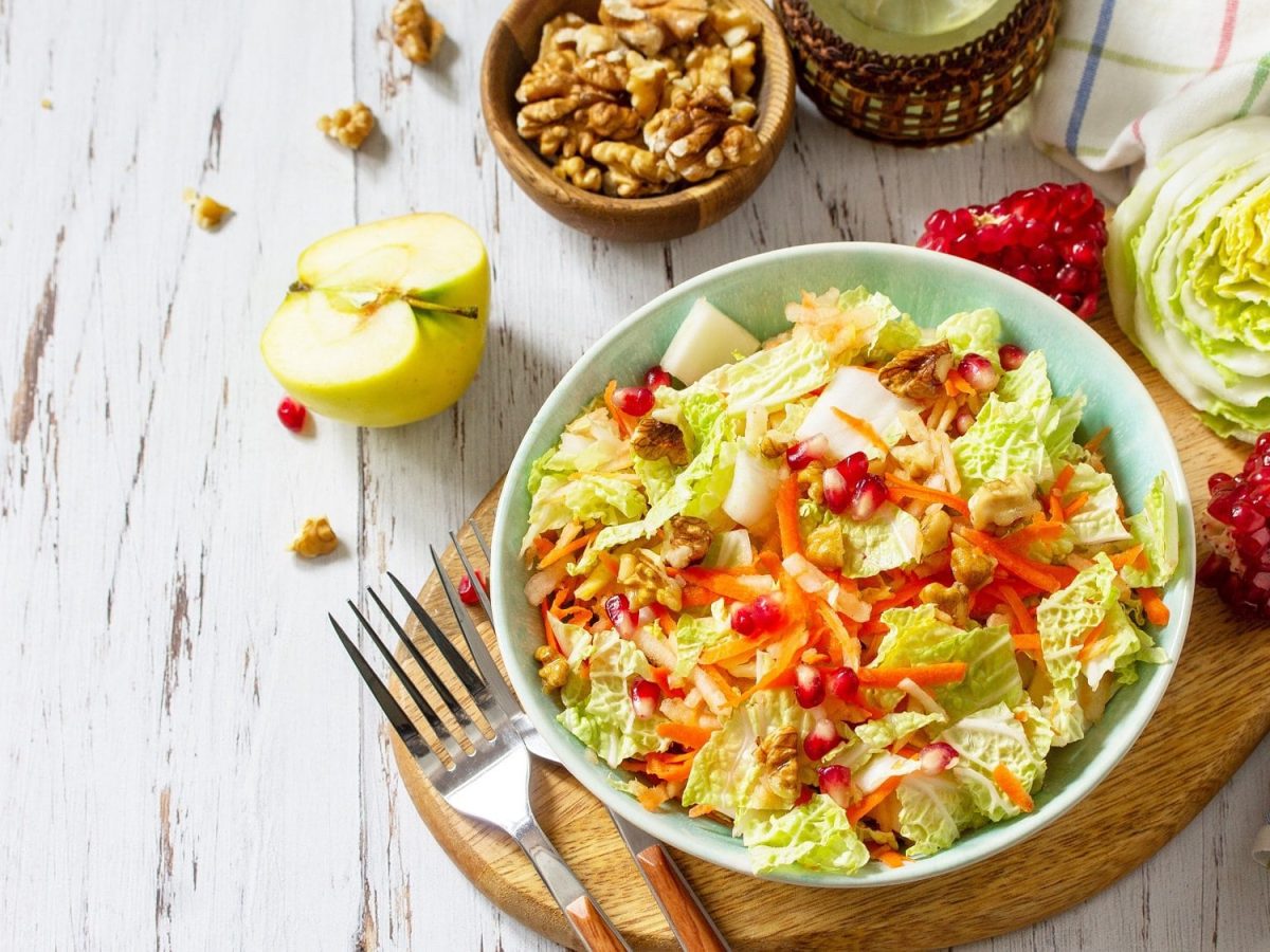 Draufsicht: Auf einem weißen Untergrund aus Holz steht eine Schüssel Chinakohl-Salat mit Granatapfelkernen. Drumherum liegen einige der Zutaten.