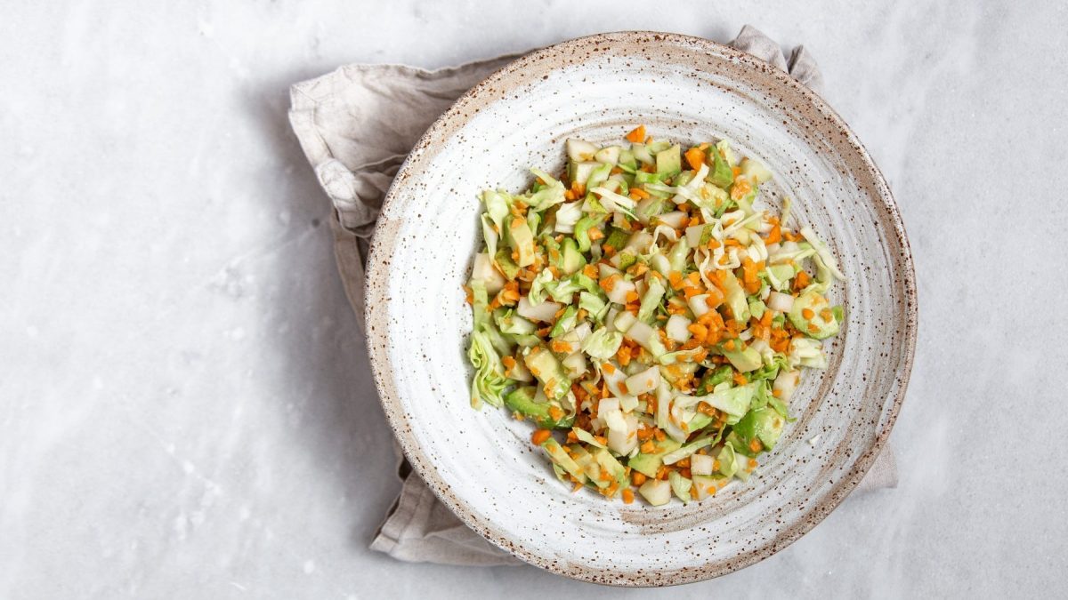 Eine graue Schale mit dem Coleslaw auf einem hellen Marmortisch mit einer beigen Serviette darunter.