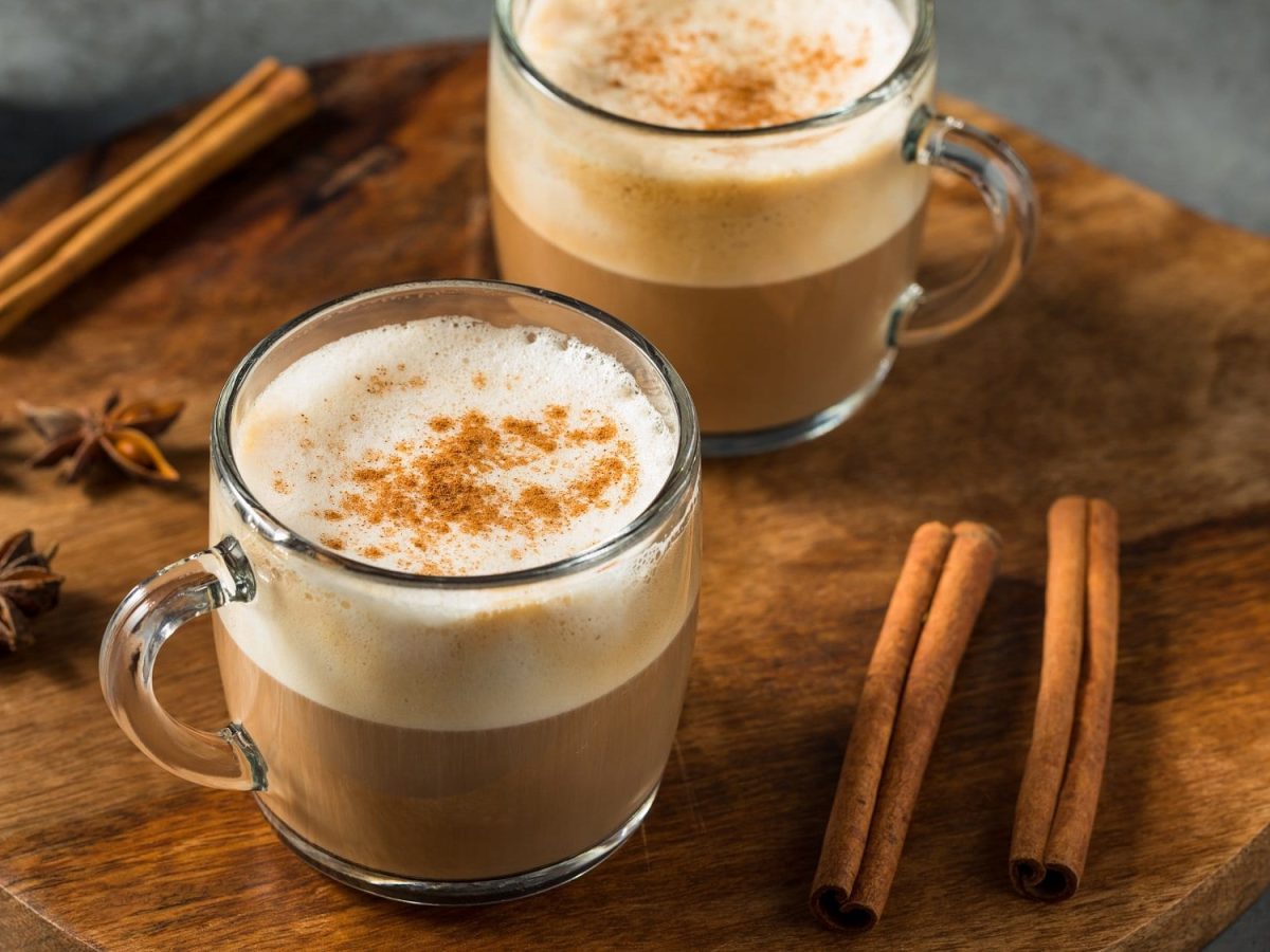 Zwei Gläser Dirty Chai Latte auf einem runden Holzbrett, daneben Zimtstangen und Anissterne.