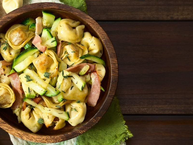 Eine Holzschüssel mit Tortellinisalat mit Pancetta und Zucchini auf einem Holztisch.