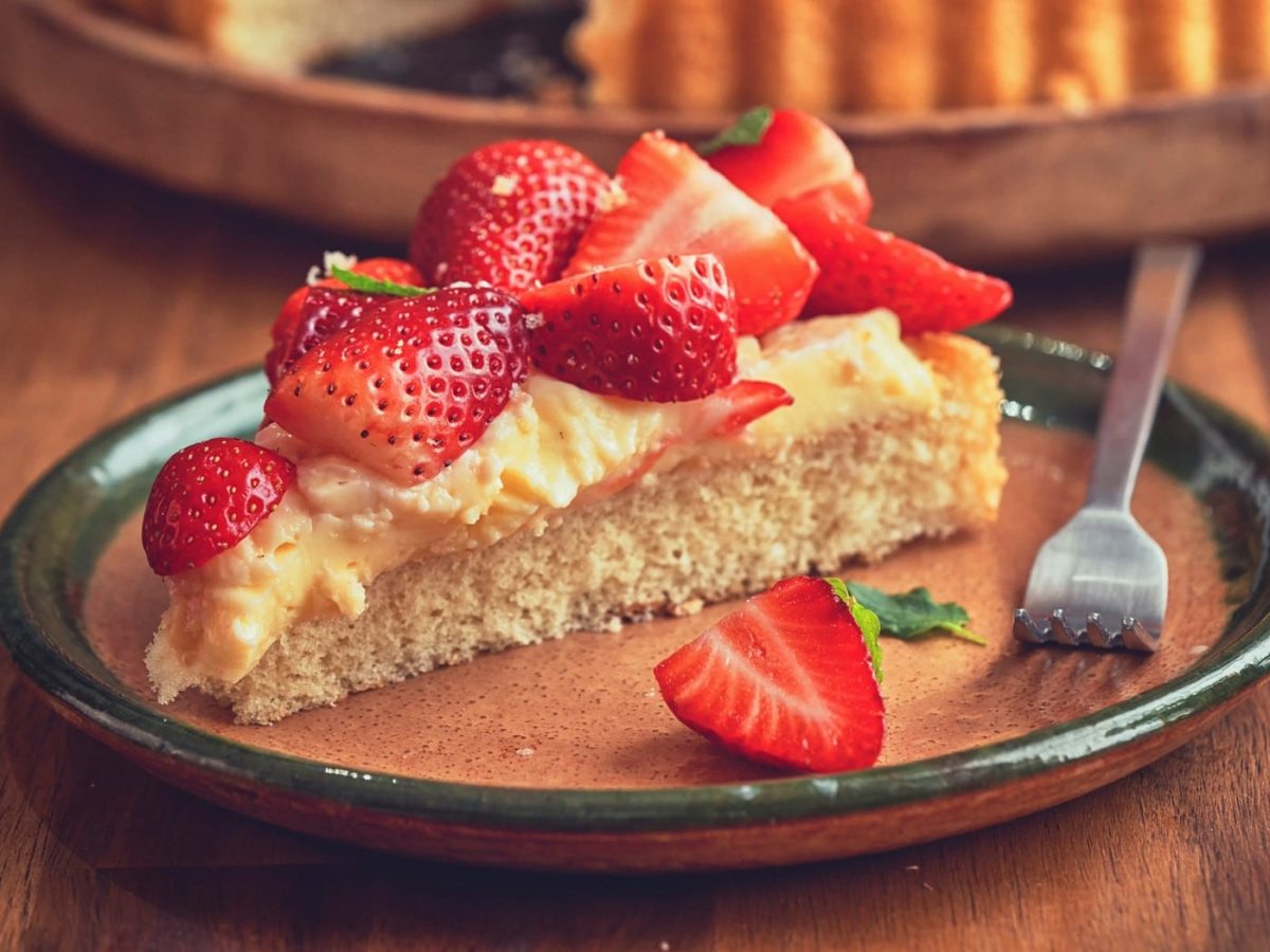 Ein Stück Erdbeer-Pudding-Kuchen mit Kuchengabel auf Teller auf Holz. Im Hintergrund angedeutet Tarte-Form. Frontalansicht.