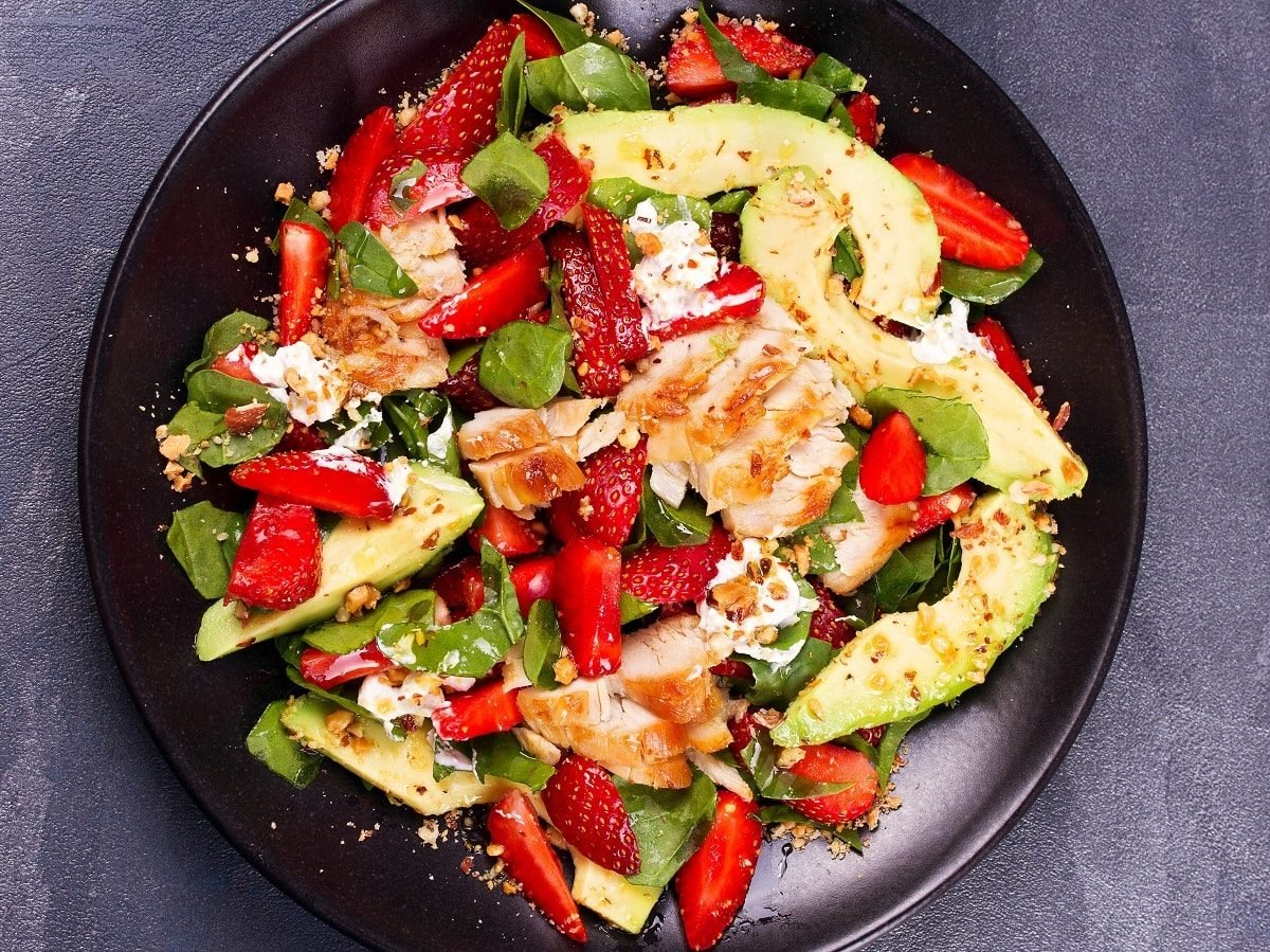 Ein schwarzer Teller mit Erdbeersalat mit Hähnchen und Feta auf einem grauen Tisch.