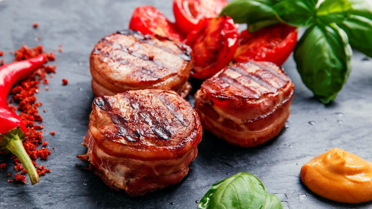 Schweinefilet im Speckmantel auf Schieferplatte. Daneben Basilikum-Blätter, gegrillte Tomaten, Chili und Sauce.