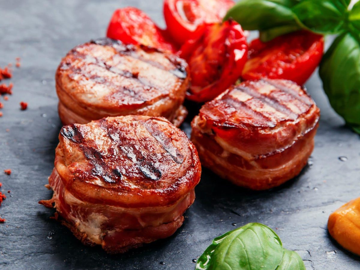 Schweinefilet im Speckmantel auf Schieferplatte. Daneben Basilikum-Blätter, gegrillte Tomaten, Chili und Sauce.