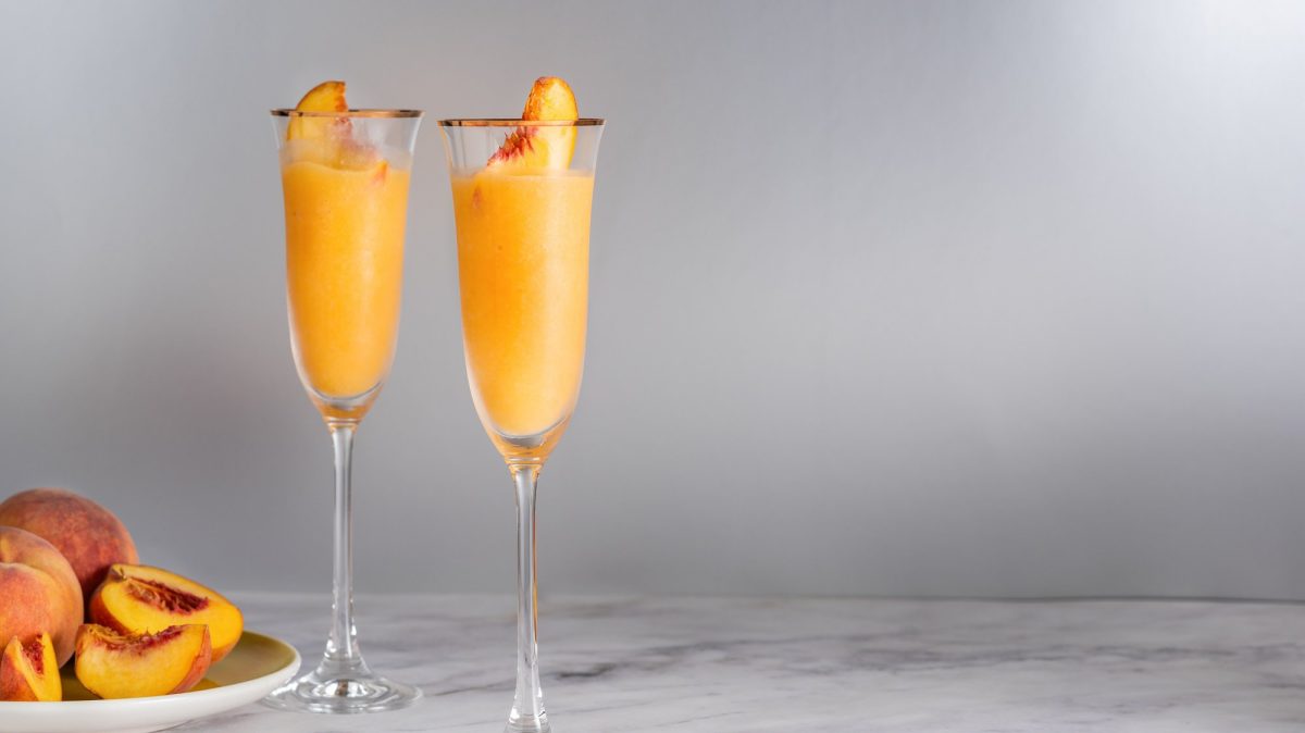 Zwei Gläser Frozen Bellini mit einer Pfirsichspalte, daneben ein Teller mit frischen, angeschnittenen Pfirsichen.