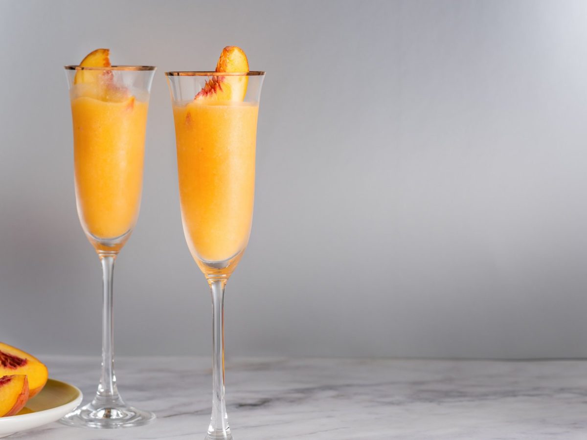 Zwei Gläser Frozen Bellini mit einer Pfirsichspalte, daneben ein Teller mit frischen, angeschnittenen Pfirsichen.