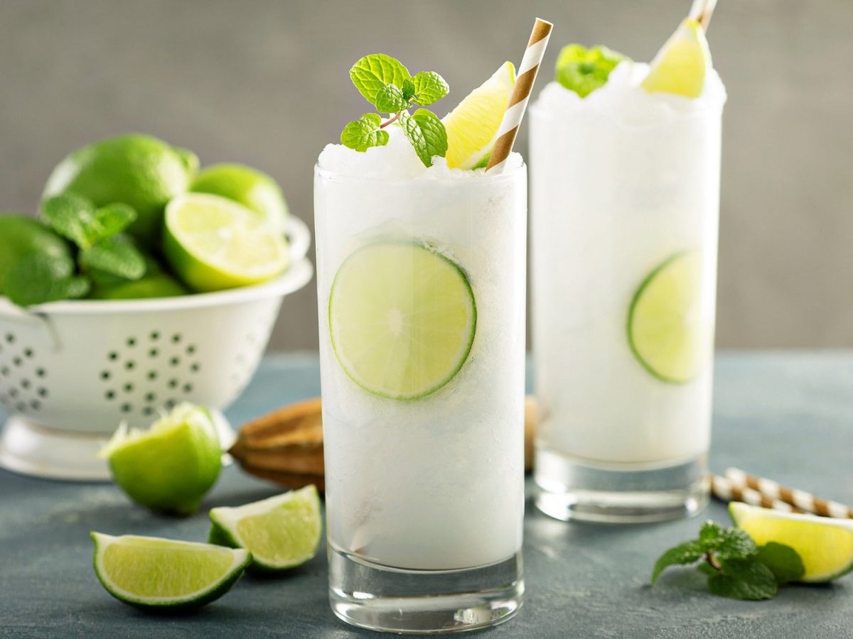 Zwei Gläser Frozen Gin Tonic mit Minze und Limettenspalten garniert, dahinter ein Sieb mit Limetten.