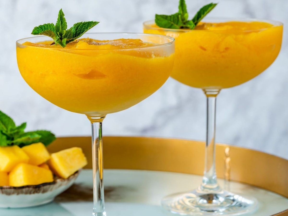 Zwei Frozen Mango Daiquiri mit Minzen auf Tablett. Im Hintergrund eine Schale mit Mangostücken und Minze. Frontalansicht.