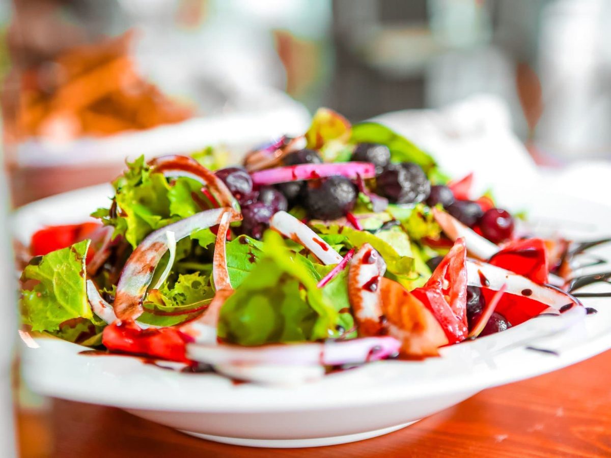 Frischer Frühlingssalat mit Tomaten, roten Zwiebeln, Oliven und Erdbeer-Vinaigrette in weißem Teller auf rotem Untergrund. Frontalansicht