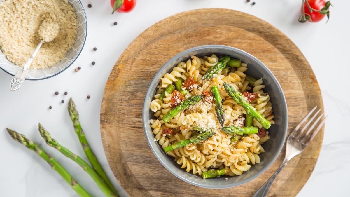 Eine graue Schüssel mit der Pasta auf einem runden Holzbrett und weißem Tisch direkt von oben fotografiert.