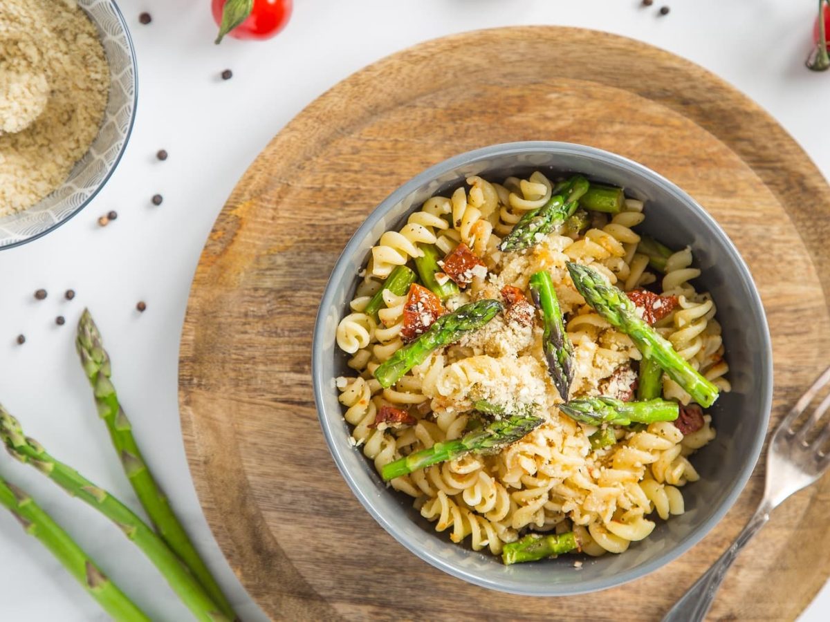 Eine graue Schüssel mit der Pasta auf einem runden Holzbrett und weißem Tisch direkt von oben fotografiert.