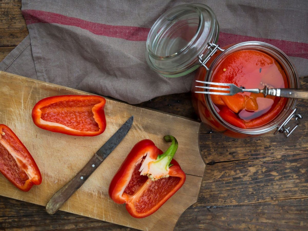 Antipasti geröstete Paprika in Olivenöl in einem Glas mit Gabel, geschnittener Paprika mit Messer auf Holzbrett und grauem Tuch auf Holz. Draufsicht.