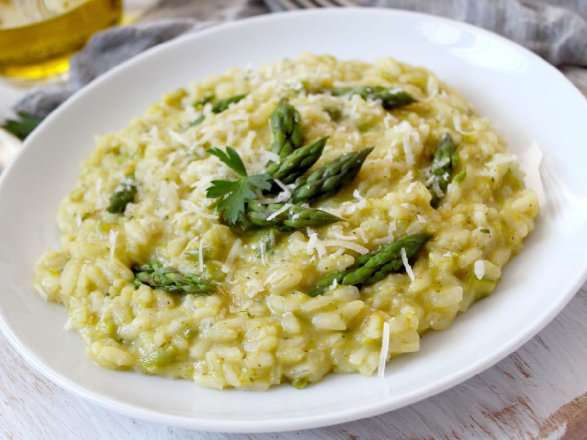 Gorgonzola-Spargel-Risotto auf weißem Teller und hellem Untergrund. Im Bild eine Schale mit Olivenöl und Parmesan auf Käsereibe. Draufsicht.