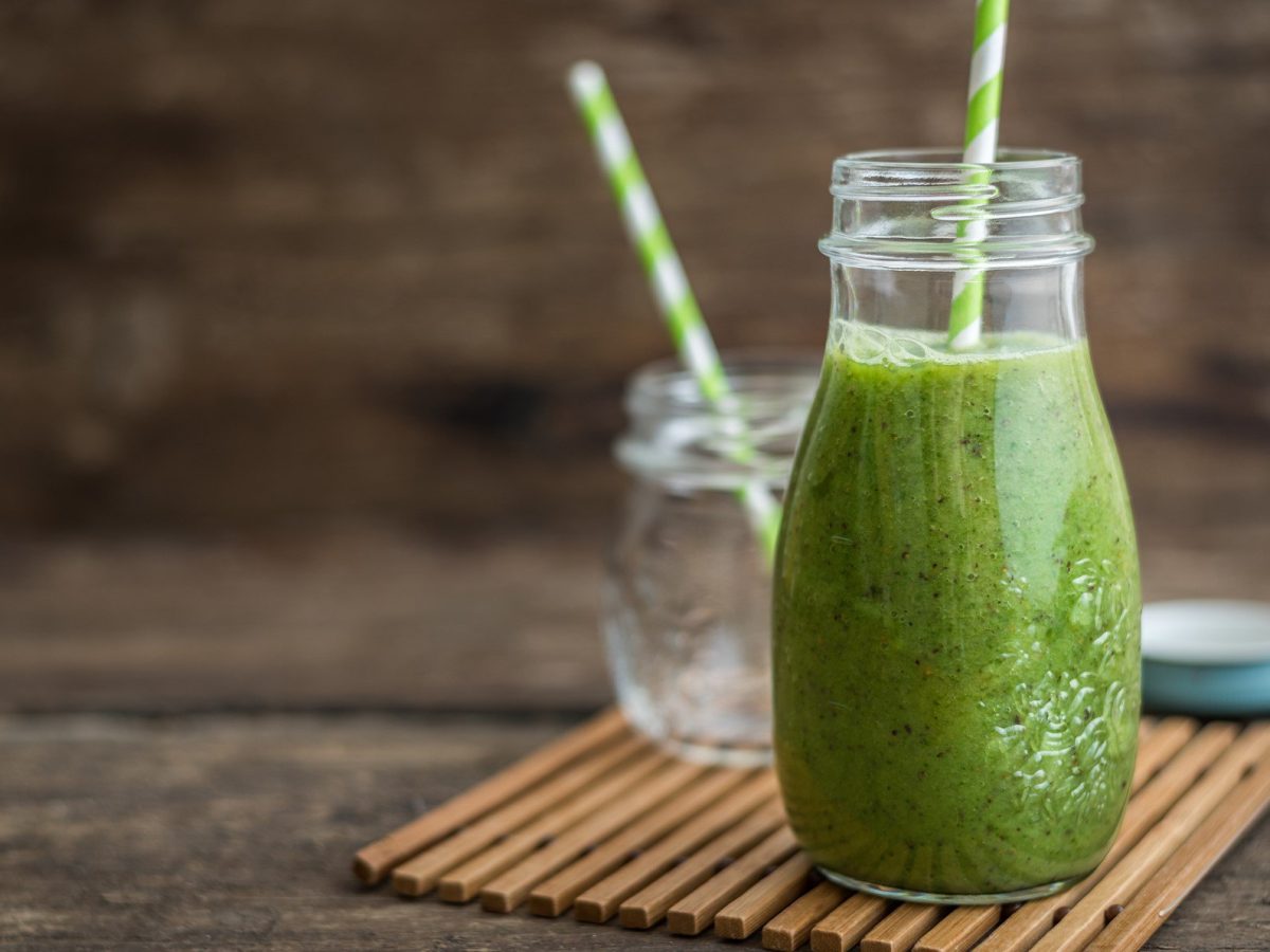 Green Goddess Smoothie im Glas mit grünem Strohhalm auf Bastmatte und Holz. Daneben leeres Glas und Schraubdeckel. Frontalansicht.