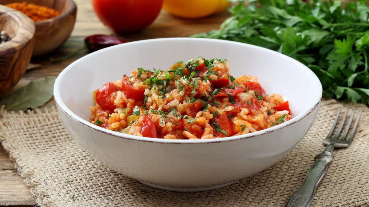 eine Schüssel griechischer Tomatenreis mit Spinat und frisch gehackter Petersilie auf einem Platzdeckchen, daneben eine Gabel, im Hintergrund frische Zutaten.