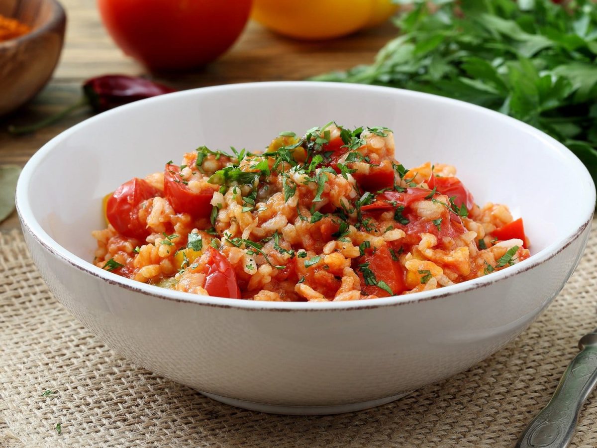 eine Schüssel griechischer Tomatenreis mit Spinat und frisch gehackter Petersilie auf einem Platzdeckchen, daneben eine Gabel, im Hintergrund frische Zutaten.