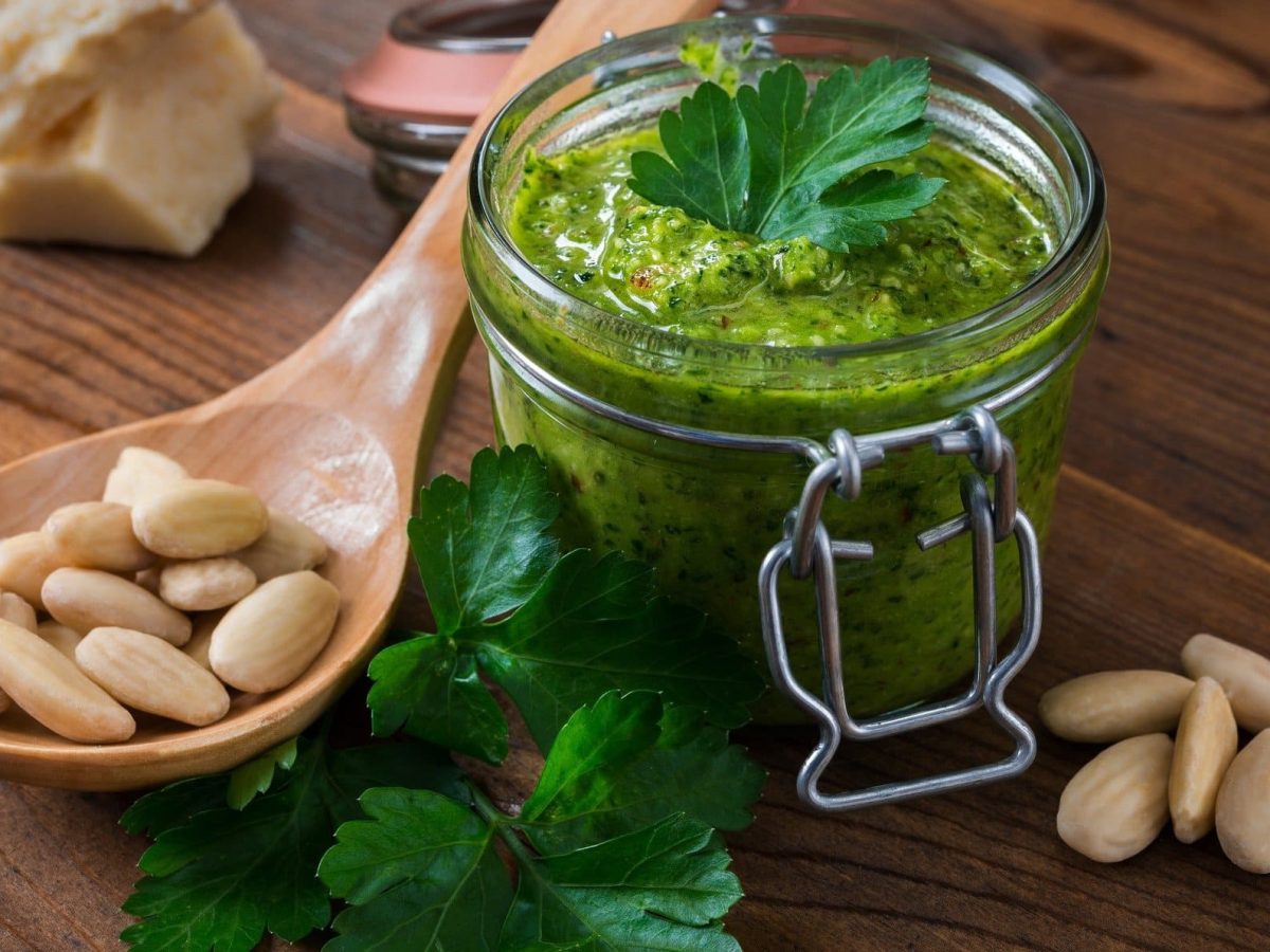 Draufsicht schräg: Auf Holz steht ein Glas Mandel-Petersilien-Pesto. Daneben liegen Mandeln ohne Haut, sowie ein Holzlöffel.