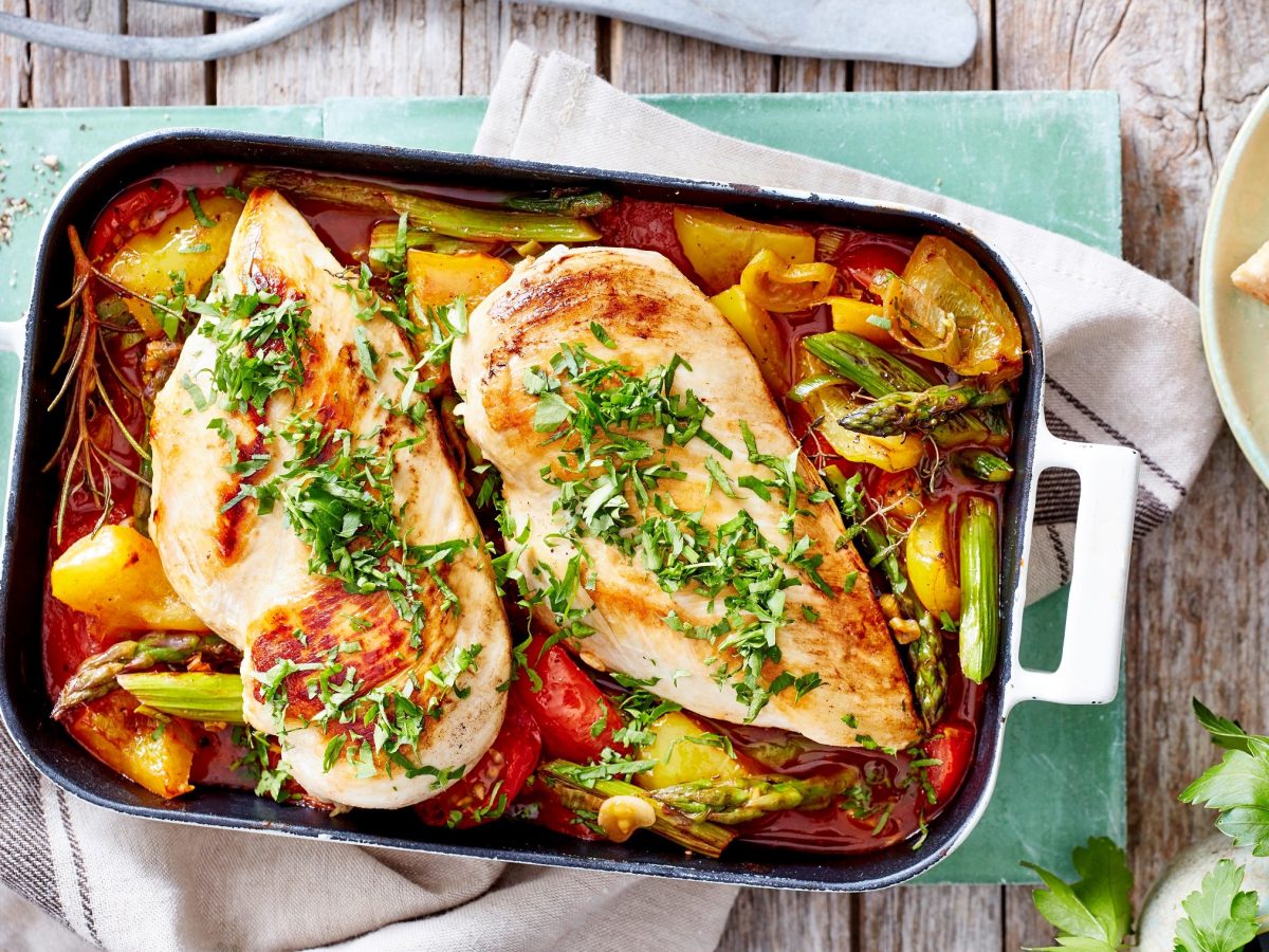 Hähnchenfilet auf Frühlingsratatouille in Auflaufform auf hellem Tuch und Holz. Im Bild eine Schale mit Baguette-Scheiben. Frische Kräuter im Bild. Draufsicht.