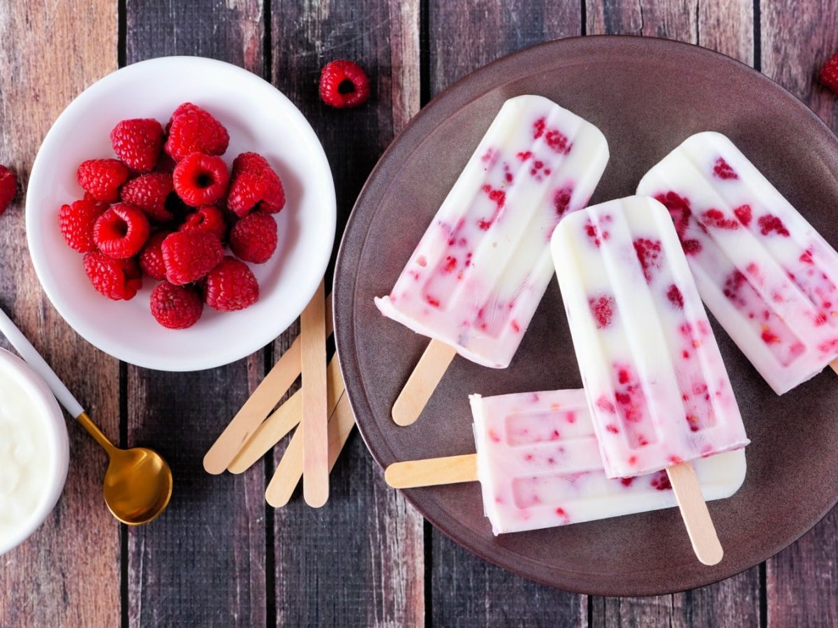 Draufsicht: Vier Himbeer-Vanille-Paletas (Eis am Stiel) auf einem braunen Teller, daneben Schale Himbeeren und Schale Joghurt mit Löffel auf Holzuntergrund.