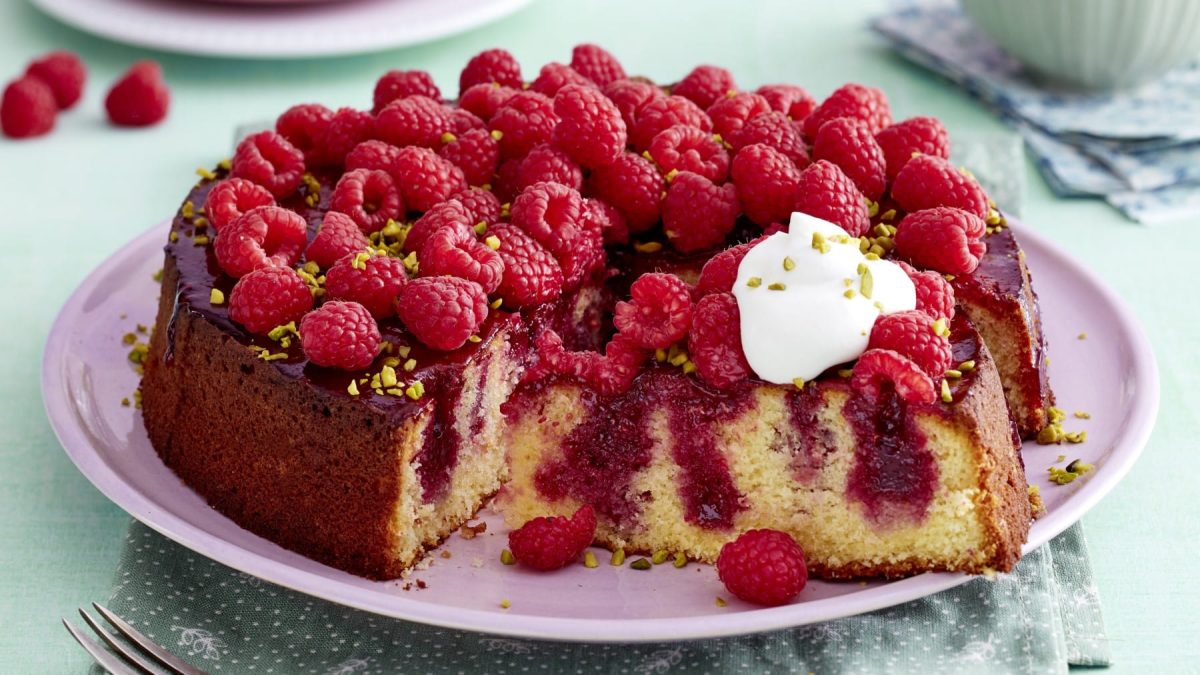 Angeschnittener Himbeer-Zitrus-Kuchen mit einem Klecks Schlagsahne auf einem rosa Teller. hellblauer Untergrund mit Geschirr und Himbeeren im Hintergrund.