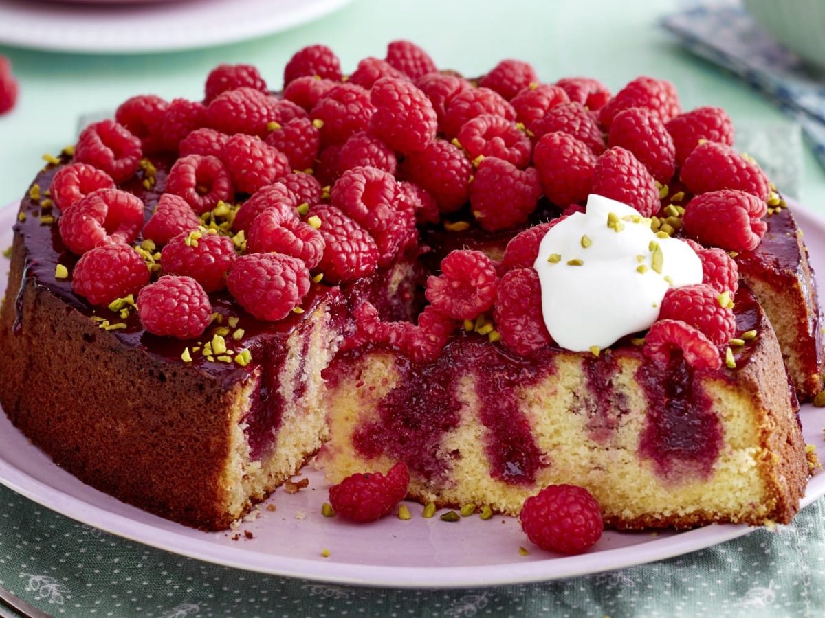 Angeschnittener Himbeer-Zitrus-Kuchen mit einem Klecks Schlagsahne auf einem rosa Teller. hellblauer Untergrund mit Geschirr und Himbeeren im Hintergrund.