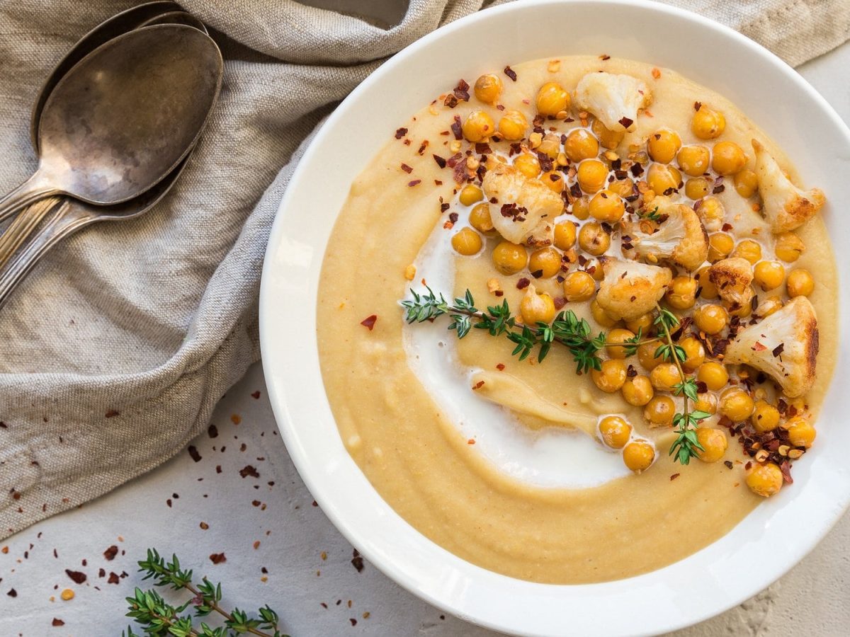 Ein Teller Hummus-Suppe mit Kichererbsen, Pfeffer und Kräutern garniert, Draufsicht, daneben zwei Löffel und Knoblauchzehen.