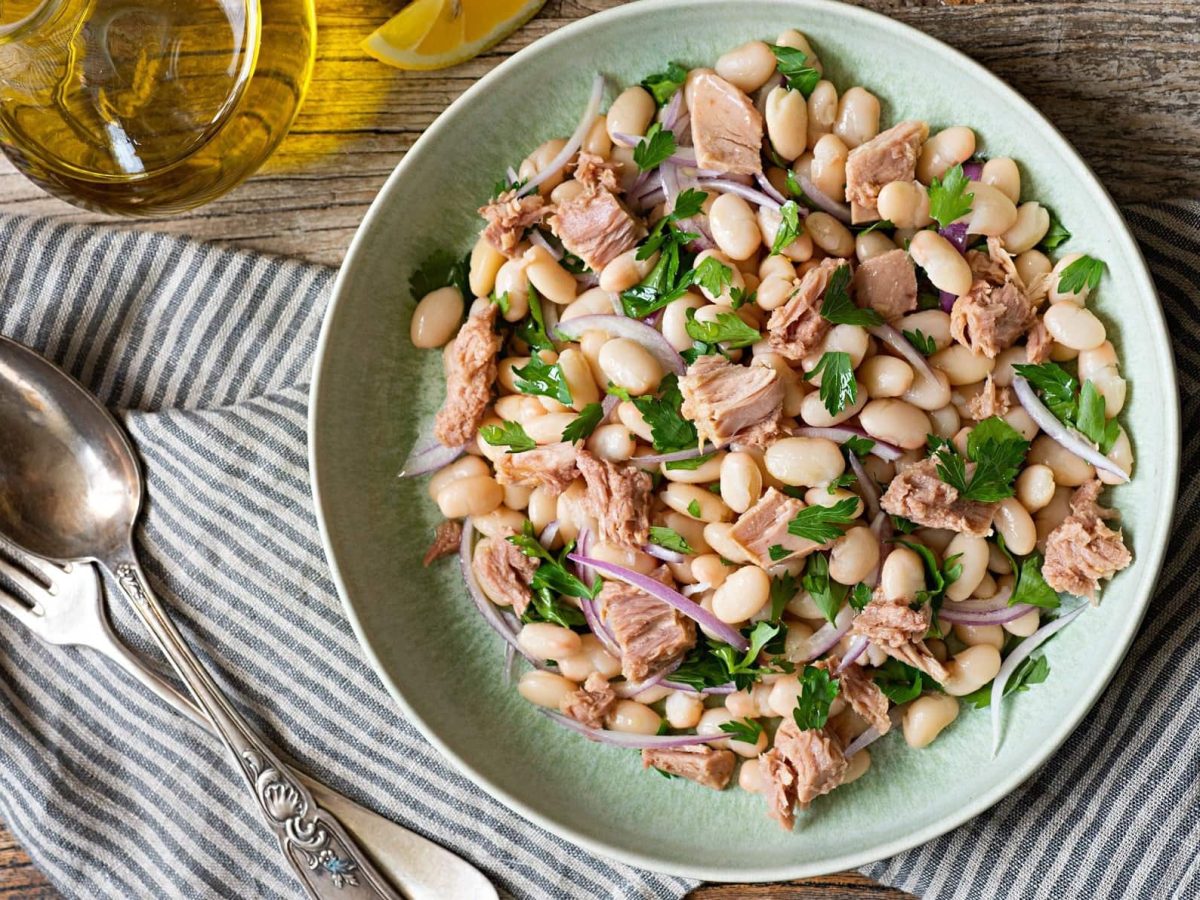 Draufsicht: Auf einem Holztisch steht ein weißer Teller Insalata di fagioli e tonno. Daneben sind ein Besteck, ein Glas mit einer ölig-gelben Flüssigkeit sowie ein Tuch.