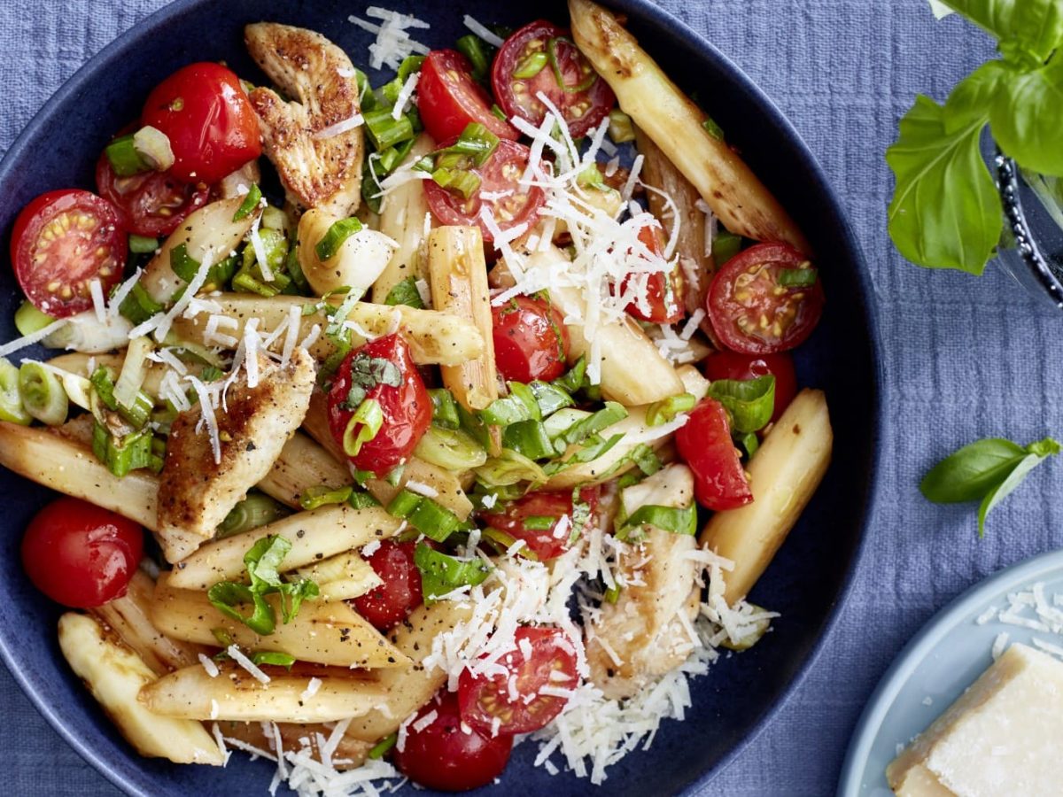 Eine dunkelblaue Bowl mit Spargel, Hähnchenfilet, Tomaten, Parmesan und Frühlingszwiebeln auf einem blauen Hintergrund. Rechts oben ein Basilikum, rechts unten Parmesan.