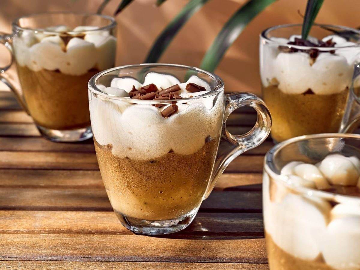 Vier Espressogläser gefüllt mit Kaffeecreme und Sahnehäubchen on top auf einem Holztisch im Halbschatten. Im Hintergrund ein paar Palmenblätter.