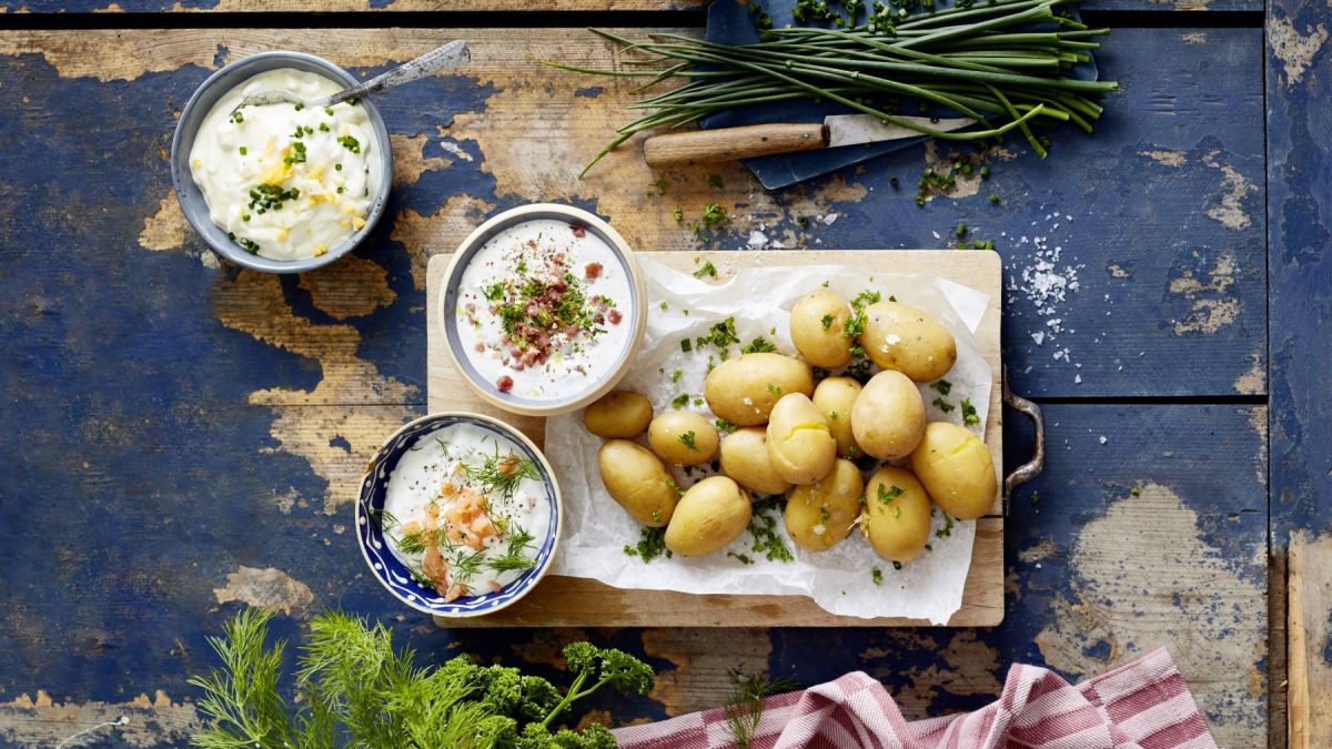 Draufsicht: Ganze Kartoffeln mit dreierlei Dip und Kräutern auf einem Brett, daneben drei Schüsseln mit verschiedenen Dips und Kräuter auf blauem Holzuntergrund.