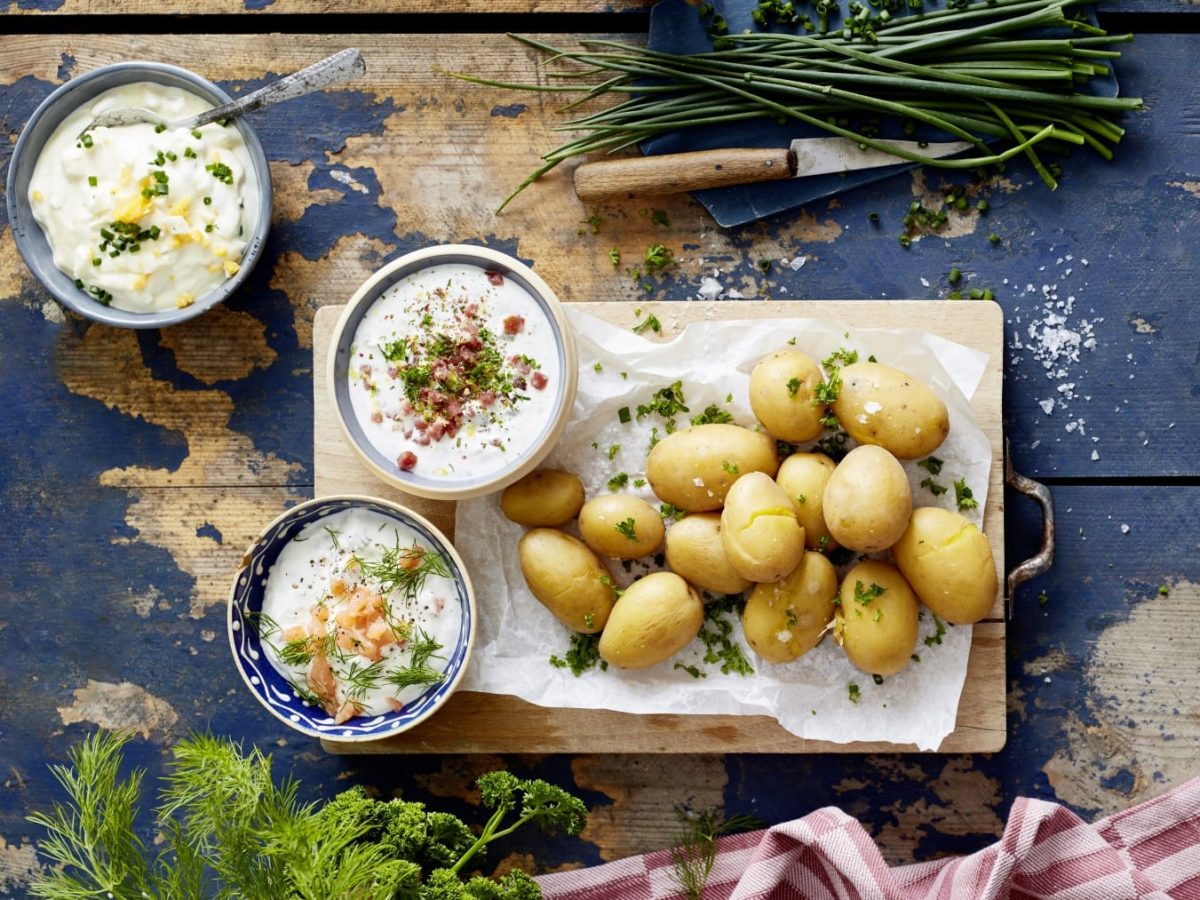 Draufsicht: Ganze Kartoffeln mit dreierlei Dip und Kräutern auf einem Brett, daneben drei Schüsseln mit verschiedenen Dips und Kräuter auf blauem Holzuntergrund.