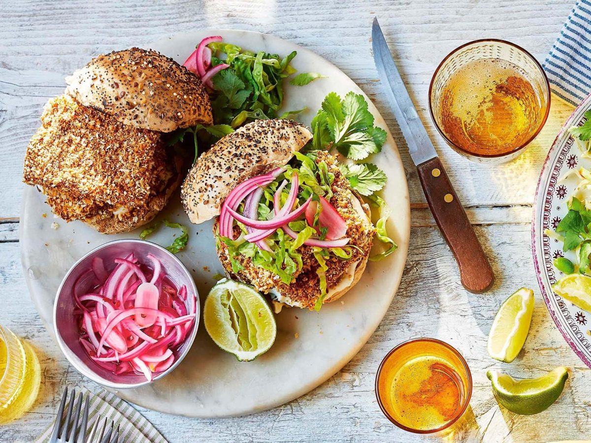 Ein gegrillter Katsu-Burger auf einem Teller mit pinken eingelegten Zwiebeln in der Draufsicht, daneben gefüllte Gläser, ein Messer und eine Schale Salat.