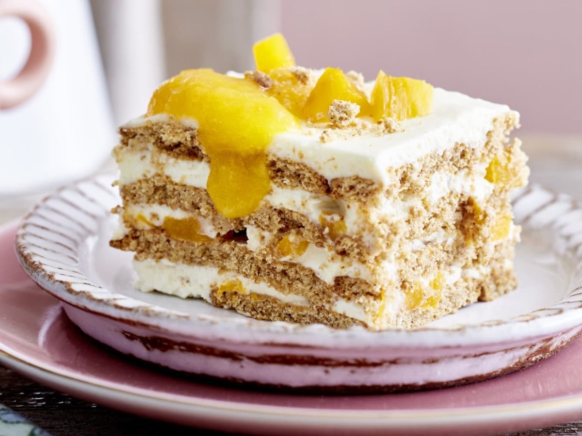 Eine Pfirsichschnitte auf einem weißen Dessertteller mit goldener Verzierung, auf einem rosa Teller, auf einem Holztisch.