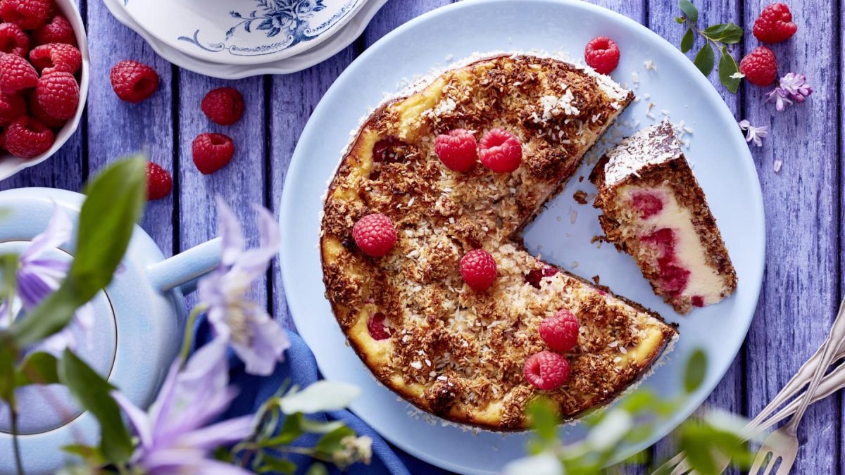 Ein Schmandkuchen mit Himbeeren auf einem hellblauen Teller auf einem hellblauen Holztisch. Auf dem Tisch sind Himbeeren, Geschirr und Besteck zu sehen.